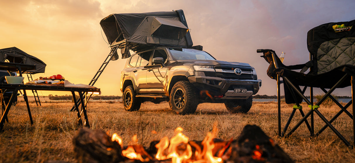 Toyota LandCruiser 300 with rooftop tent parked in a field at sunset, with a campfire in the foreground and camping chairs and a table nearby.